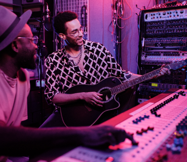 Recording instruments Two men: one operates a mixing console, the other plays guitar.